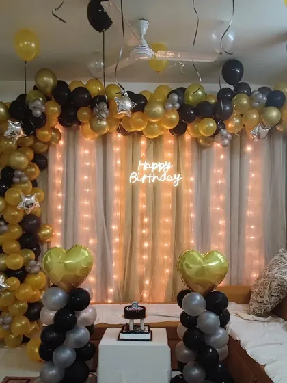 An elegant black and gold balloon garland for a birthday celebration. This setup includes a 'Happy Birthday' neon sign and a fairy light curtain, creating a sophisticated photo backdrop in the living room.