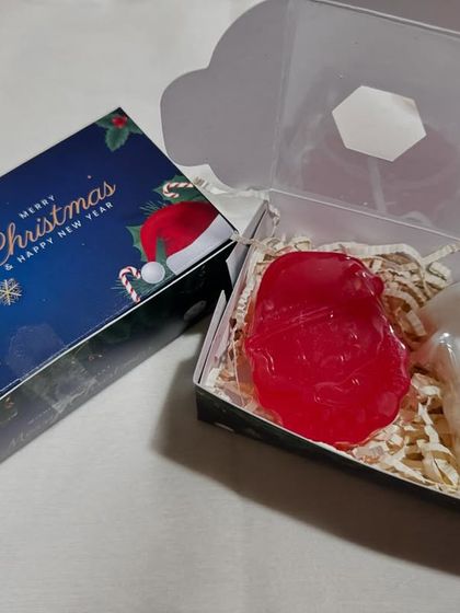 A festive Christmas gift box containing two handmade soaps, a red gem-like soap and a white Santa soap, nestled in paper shred. A simple and thoughtful holiday gift.