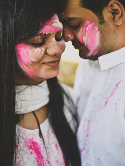 An intimate pre-wedding or post-Holi photoshoot moment. The couple is close together, their faces smeared with pink color, creating a playful and romantic image.