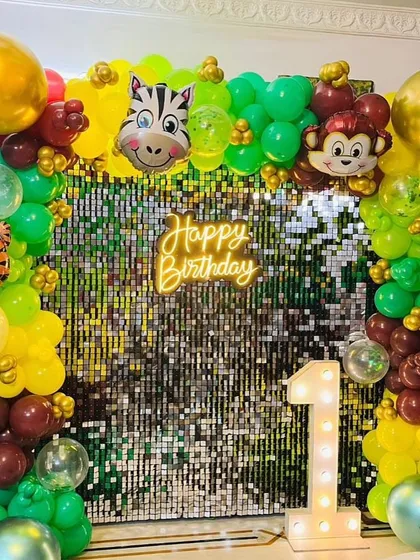 A vibrant jungle-themed first birthday party backdrop. This setup includes a shimmering sequin wall, a custom neon sign, marquee number, and a lush balloon garland with fun animal characters.