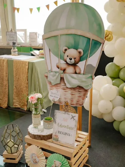 A decorative setup featuring a teddy bear in a hot air balloon basket. The "The Adventure Begins" sign and rustic props tie all the elements of the travel theme together.