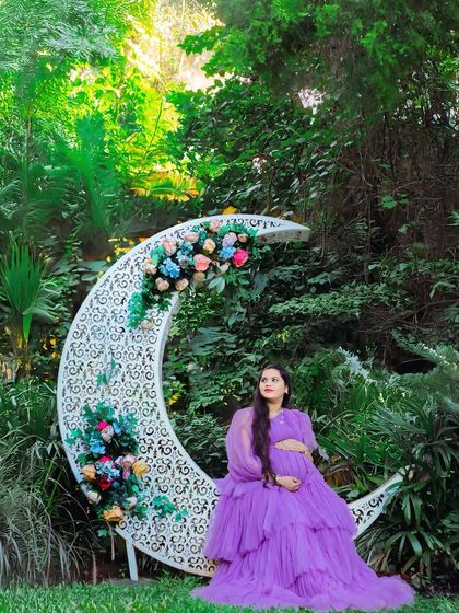 A magical outdoor portrait featuring a mother-to-be in a purple gown, seated on a large, decorative moon prop. The lush garden setting adds to the enchanting, fairytale feel of the shoot.