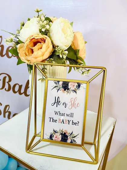 A close up of a framed 'He or She' sign, part of a gender reveal decor package. The modern gold geometric frame and floral design add a touch of elegance to the celebration.
