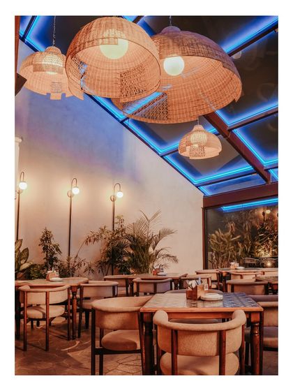 As evening approaches, the same space transforms. The skylight is illuminated with blue neon lights, creating a vibrant and energetic mood that contrasts beautifully with the warm glow of the rattan pendants.