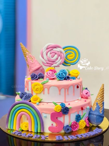 A two-tier candy world cake with a rainbow, lollipops, and ice cream cones. The pink drip and colorful flowers add to the sweet and playful theme.