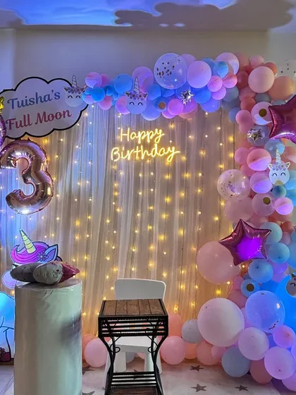 This cozy unicorn-themed decor is perfect for a house party. It includes a "Happy Birthday" neon sign against a fairy-light curtain, a number '3' balloon, and unicorn cutouts.