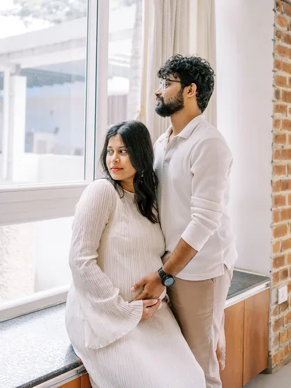 A quiet, shared moment by the window. They are looking out together, perhaps dreaming of the future with their new baby. The natural light is soft and beautiful.