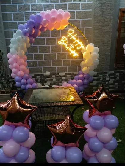 An outdoor birthday celebration at dusk. A circular balloon arch in sunset-inspired pastel colors is lit by a neon sign, with star-shaped balloon columns adding to the charm.