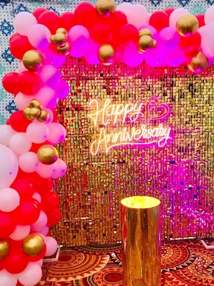 A vibrant anniversary photo booth with a gold sequin backdrop and a red, pink, and gold balloon arch. The "Happy Anniversary" neon sign adds a warm, celebratory glow to the setup.