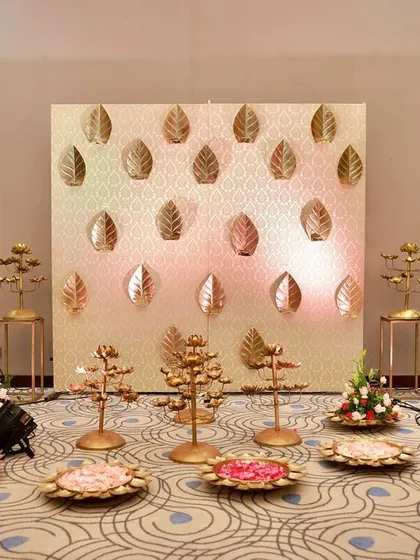 A minimalist setup for a traditional ceremony, featuring a golden leaf backdrop and ornate brass candelabras and offering plates.