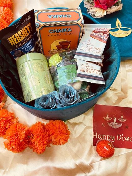 An open view of our round velvet Diwali box, filled with an assortment of teas, chocolates, and almond brittle, complemented by delicate blue paper flowers.