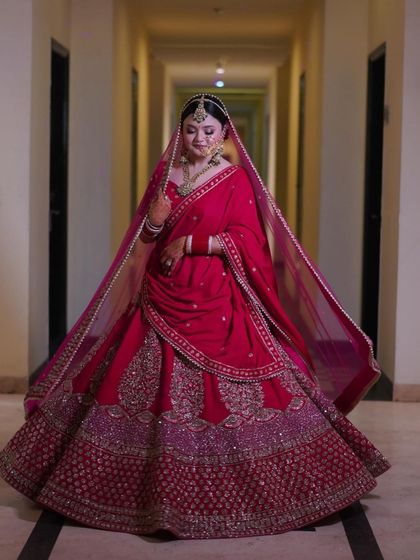 A full-length portrait of a bride in a magnificent red and maroon lehenga, standing in a brightly lit corridor.