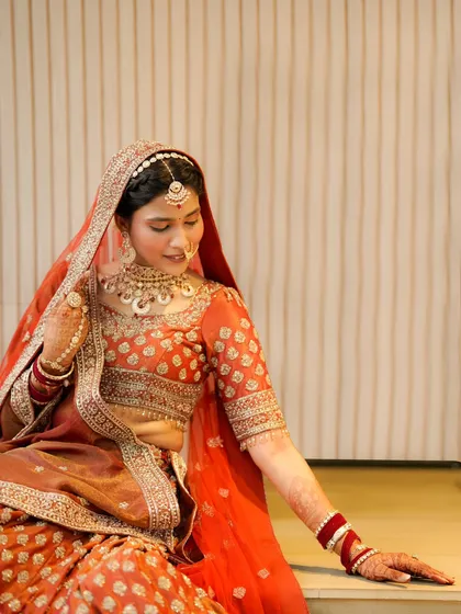 A graceful seated portrait of the bride. We focus on her gentle expression and the rich details of her orange and red lehenga.