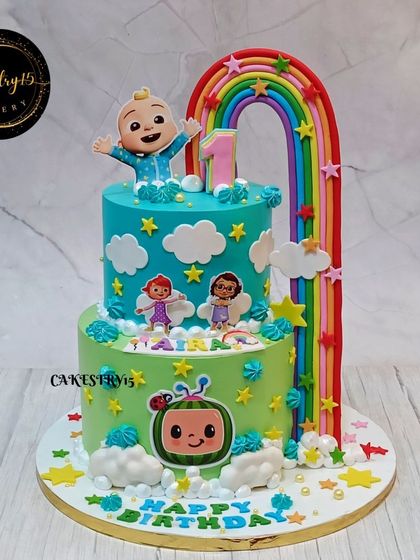 A cheerful two-tier Cocomelon cake featuring a large fondant rainbow arching over the top. The cake is decorated with edible characters and fluffy fondant clouds.