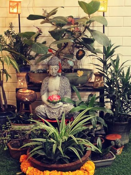 A Buddha statue surrounded by a variety of indoor and outdoor plants creates a welcoming and peaceful entrance. The arrangement is finished with marigold flowers for a traditional, auspicious touch.