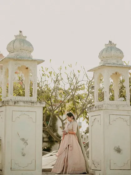 A portrait that feels like a painting. The bride stands between two traditional structures, her pastel lehenga complementing the soft light and serene architecture.