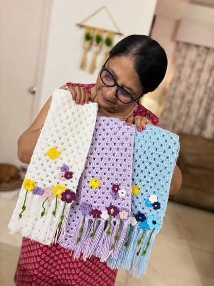 Holding up three of my floral crochet scarves in white, lavender, and light blue. It takes nearly half a day just to complete the flowers for one scarf.