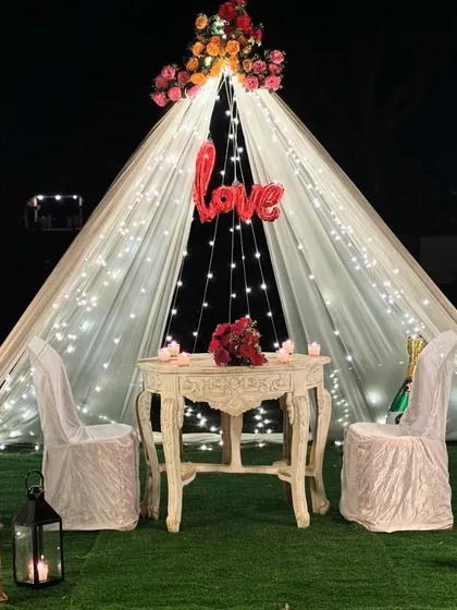An intimate and romantic proposal setup in a garden, featuring a beautiful canopy tent draped in fairy lights, a 'Love' balloon, and a candlelit table for two.