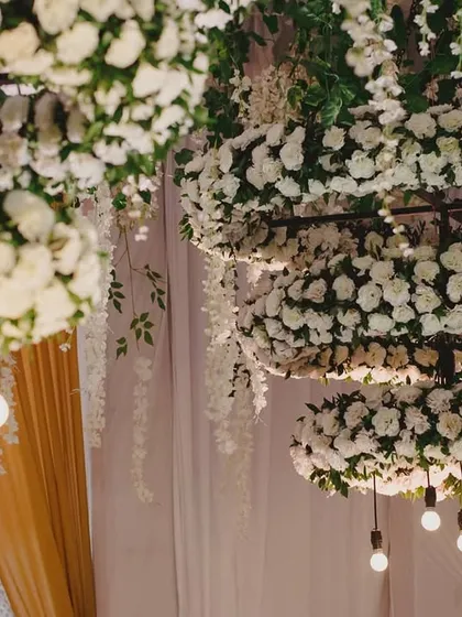 Detail shot of the floral chandeliers. The combination of dense carnation rings and exposed Edison bulbs offers a beautiful contrast between classic floral artistry and modern industrial lighting design, perfect for an evening event.