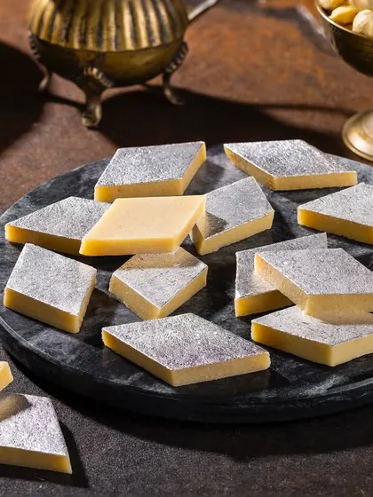 Classic Kaju Katli for Grover Sweets. The macro focus and dark marble platter emphasize the smooth texture and silver leaf (vark) of this popular Indian sweet.