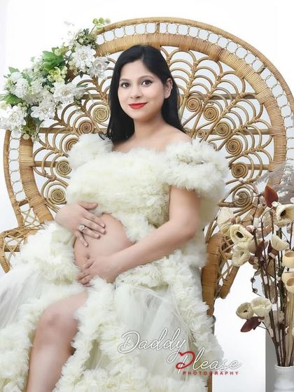 A lovely portrait of an expectant mother in a white ruffled gown, seated on an elegant peacock chair. The neutral-toned floral decor adds a touch of rustic charm to the studio setup.