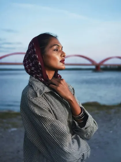 A colorful outdoor fashion portrait taken at dusk. The model, wearing a headscarf, stands by the water with a red bridge in the background, showcasing a blend of fashion and environmental portraiture.