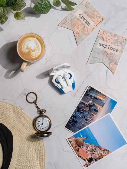 A travel-themed flat-lay for boAt Airdopes. The earbuds are styled with a pocket watch, postcards, and a sun hat, suggesting they are the perfect companion for any adventure.