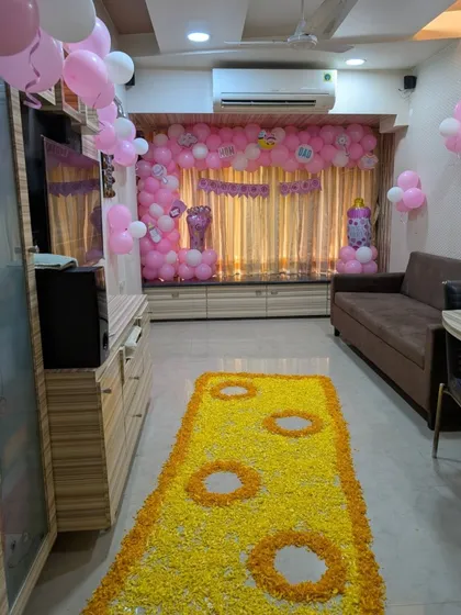 A full view of a living room decorated for a baby girl's welcome. The pink balloon backdrop is complemented by a long yellow floral runner, designed to look like a traditional 'paaypudi' or foot mat.