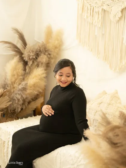 A happy and candid moment of a mother-to-be in a black dress. She is seated in a boho-styled room, smiling down at her baby bump with pure joy.