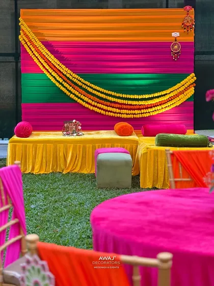 A traditional Mehendi seating area, with a colorful backdrop of orange, pink, and green drapes, and low seating with bolsters.