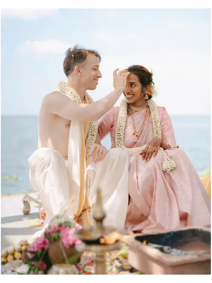 A tender moment during a seaside Hindu ceremony, as the groom applies a tilak to the bride's forehead. The sea behind them adds a sense of infinite possibility to their union.