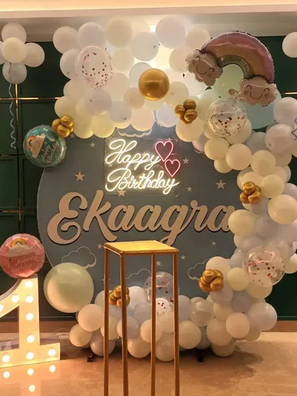 A dreamy rainbow-themed first birthday for "Ekaagra." This setup includes a personalized circular backdrop, a white and gold balloon arch, a "Happy Birthday" neon sign, and a light-up number "1."