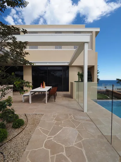 The outdoor dining and patio area of a coastal home, featuring crazy paving and a simple pergola. The design prioritizes the view and creates a functional space for enjoying the sea breeze.