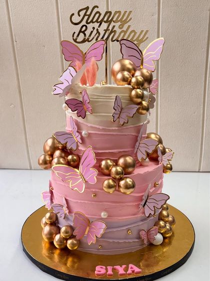 Fluttering into celebration with this dreamy two-tiered butterfly birthday cake. The design features shades of pink buttercream, delicate butterflies, and golden spheres for a touch of glam.