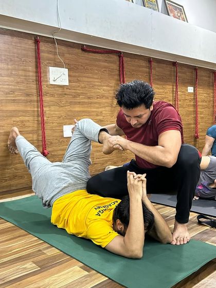 An instructor provides a gentle yet firm adjustment in Supta Virasana (Reclining Hero Pose) to help a student open their quadriceps and hips.