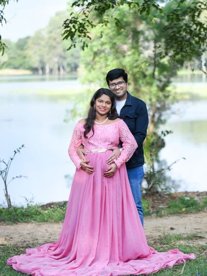 A smiling couple portrait by the lake, with the mom-to-be in a pink lace dress.