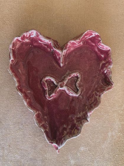 A heart-shaped trinket dish with a bow detail, glazed in a rich magenta. This piece was hand-built and is perfect for holding your most treasured small items.