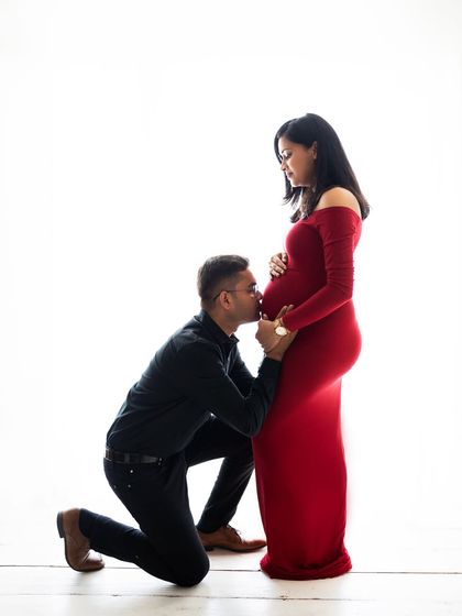 A classic and elegant maternity portrait. The dad-to-be kisses his partner's baby bump while she wears a striking red gown. A timeless image of love and anticipation.