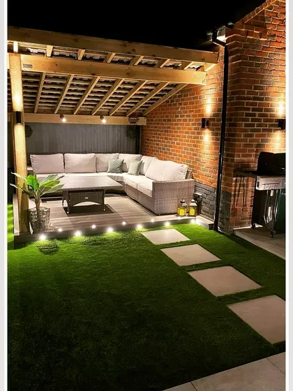 A cozy backyard corner featuring a wooden pergola attached to a brick wall. The space is transformed into an intimate lounge with a large sectional sofa and ambient lighting from string lights and ground-level spotlights.