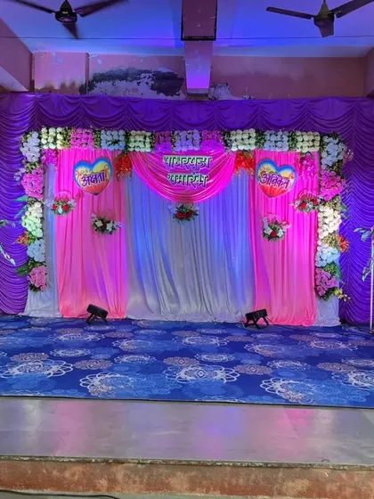 A colorful stage setup for a wedding reception or sangeet. The purple and pink drapes are decorated with floral arrangements and uplighting.