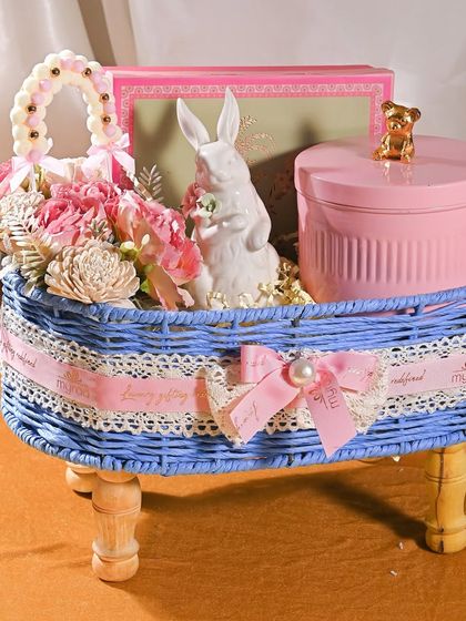 A whimsical baby announcement hamper in a blue wicker basket. It includes a ceramic bunny keepsake, a pink canister with a teddy bear topper, and a delicate floral arrangement.