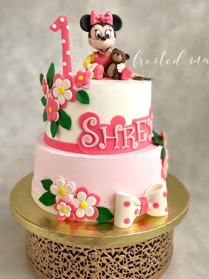 A sweet two-tiered cake for a first birthday, featuring a handmade baby Minnie Mouse figurine with her teddy bear. The soft pink colors and delicate flowers make it an adorable centerpiece.