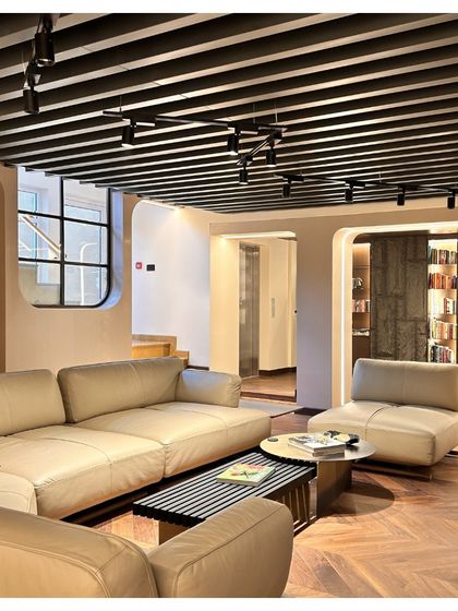 A wider view of the study lounge, showing how the space opens up while maintaining its cozy feel. The herringbone pattern of the wooden floor adds a classic touch, contrasting with the modern slatted ceiling and track lighting.