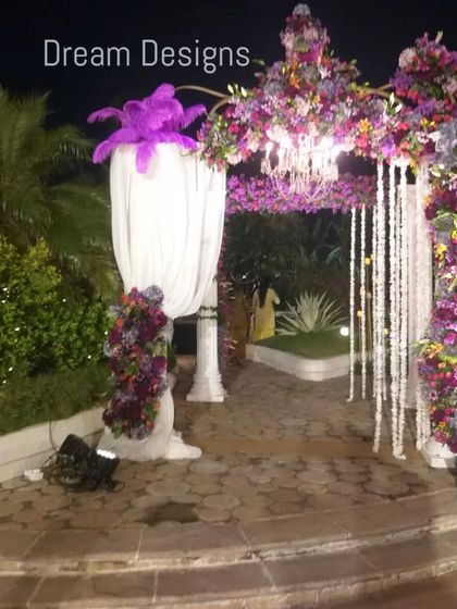 A walkway to remember. This unique entrance features pillars draped in white fabric and crowned with dramatic purple floral arrangements, with a floral arch connecting them to create a regal and memorable pathway.