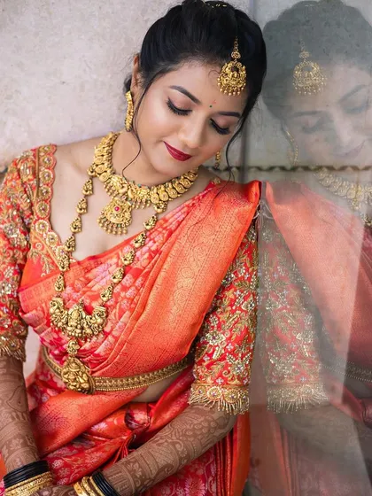 This is an artistic bridal portrait that captures the soft, glowing makeup beautifully. The elegant pose and the reflection highlight the flawless finish and the classic red lip, perfect for a South Indian wedding.