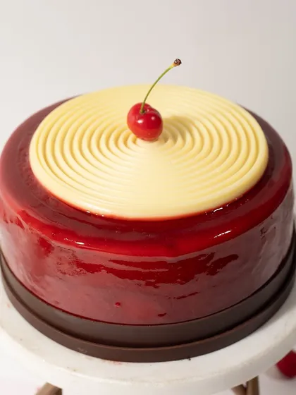 A sleek and modern cake with a glossy red mirror glaze, a white chocolate spiral, and a single cherry on top. It's minimalist, elegant, and delicious.