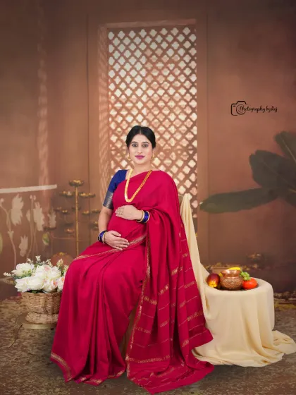 A regal studio portrait of a mother-to-be in a red saree, seated gracefully with traditional props.