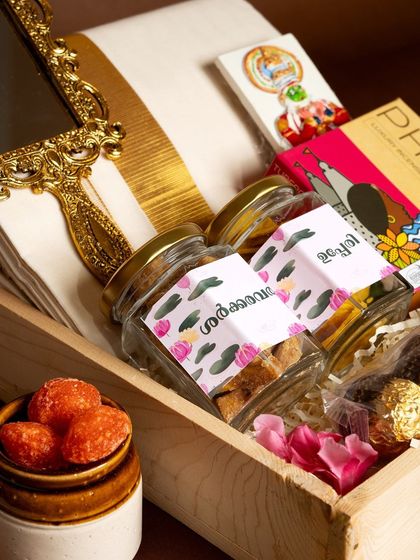 A close-up of the 'Pichi Poo' hamper, highlighting the jars of upperi and sharkara varatti with custom labels in Malayalam, alongside a traditional Aranmula kannadi (mirror).