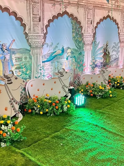 Another angle of the Pichwai-themed decor, focusing on the detailed artwork on the wall panels. The scene depicts Krishna and peacocks in a lush landscape, enhanced by the foreground arrangement of cow cutouts and marigolds.