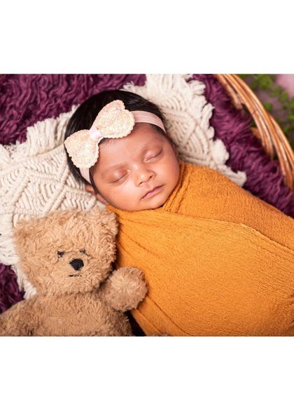 A close-up of a sleeping baby nestled in a basket with a teddy bear friend. The soft textures of the wrap and the bear create a cozy and comforting newborn portrait.